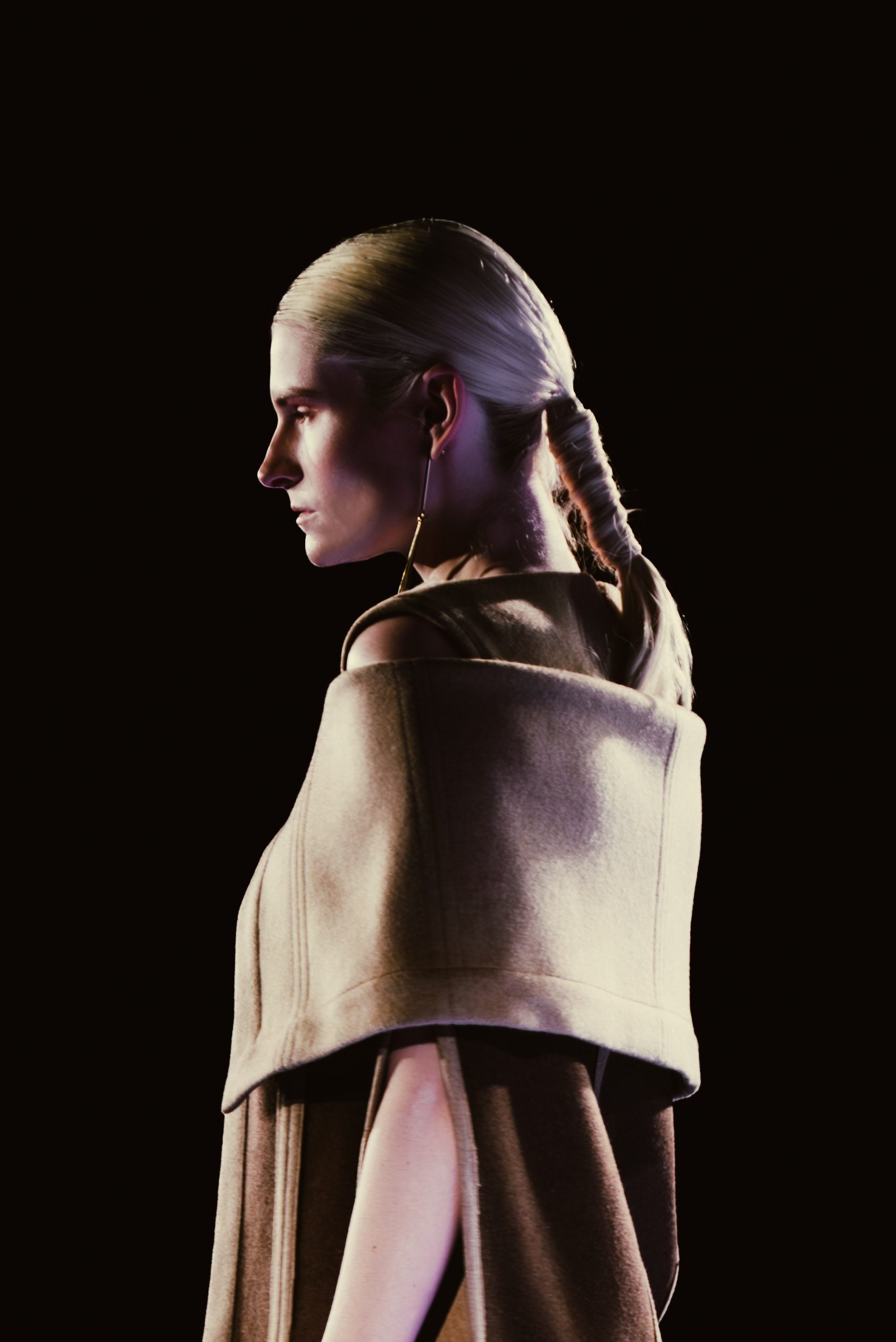 A model in a sculptural beige hood stands in profile against darkness.