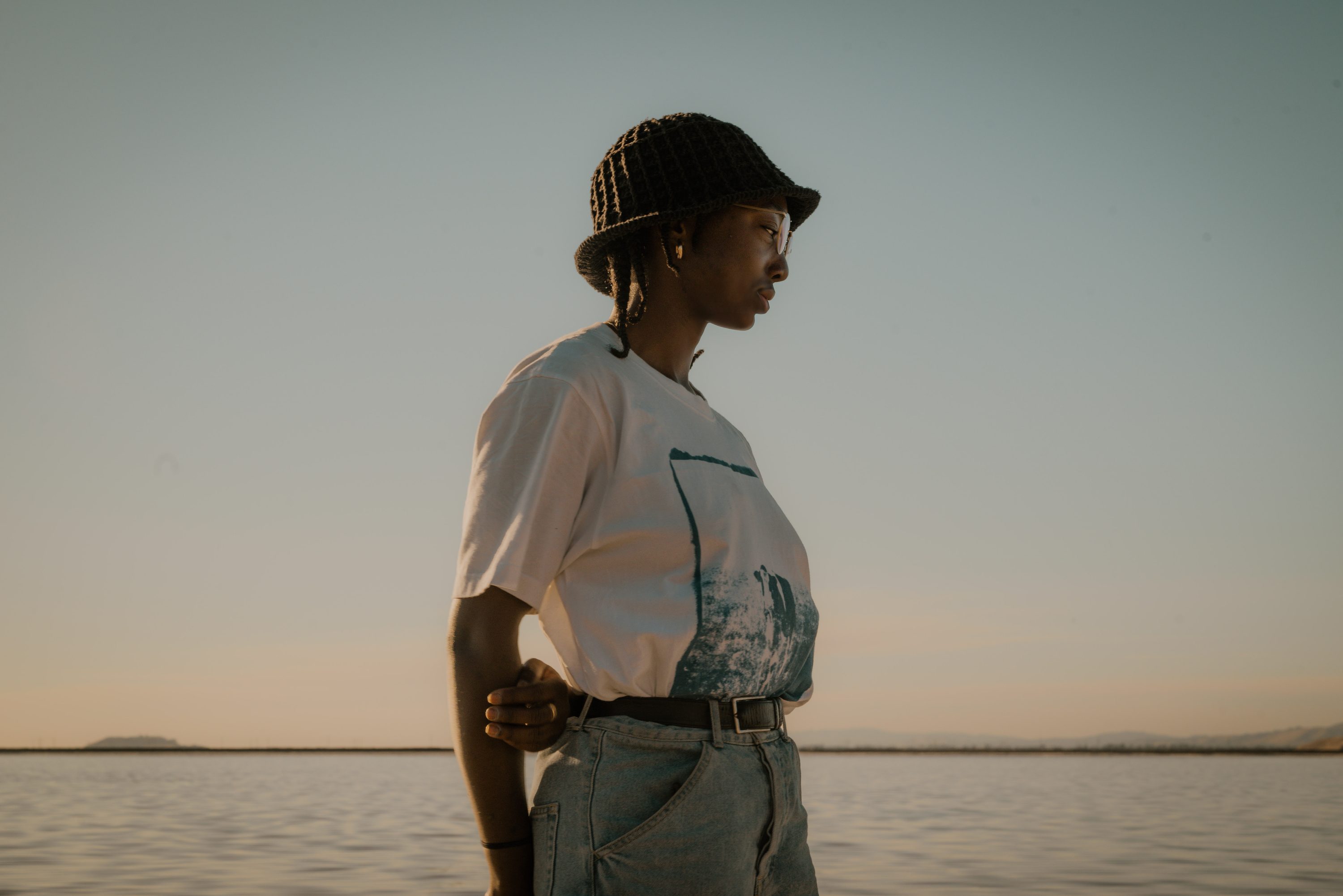 A woman in a white tee stands at the shoreline in golden-hour light.